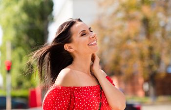 Woman in a red off-shoulder top smiles with eyes closed, standing outdoors with greenery and a blurry background.