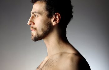 Profile view of a shirtless man with short hair and a beard against a gradient background.