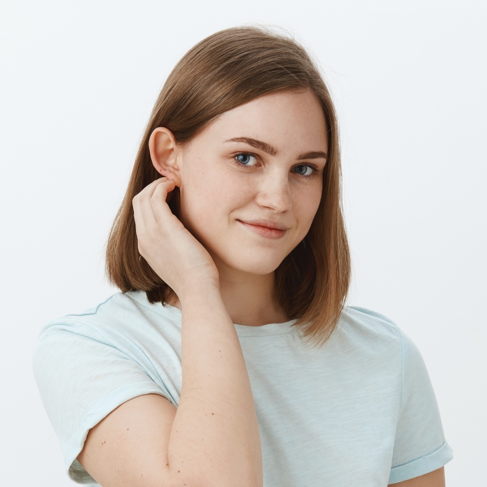 A person with shoulder-length brown hair and a light blue shirt gently touches their ear, looking into the camera against a plain background.