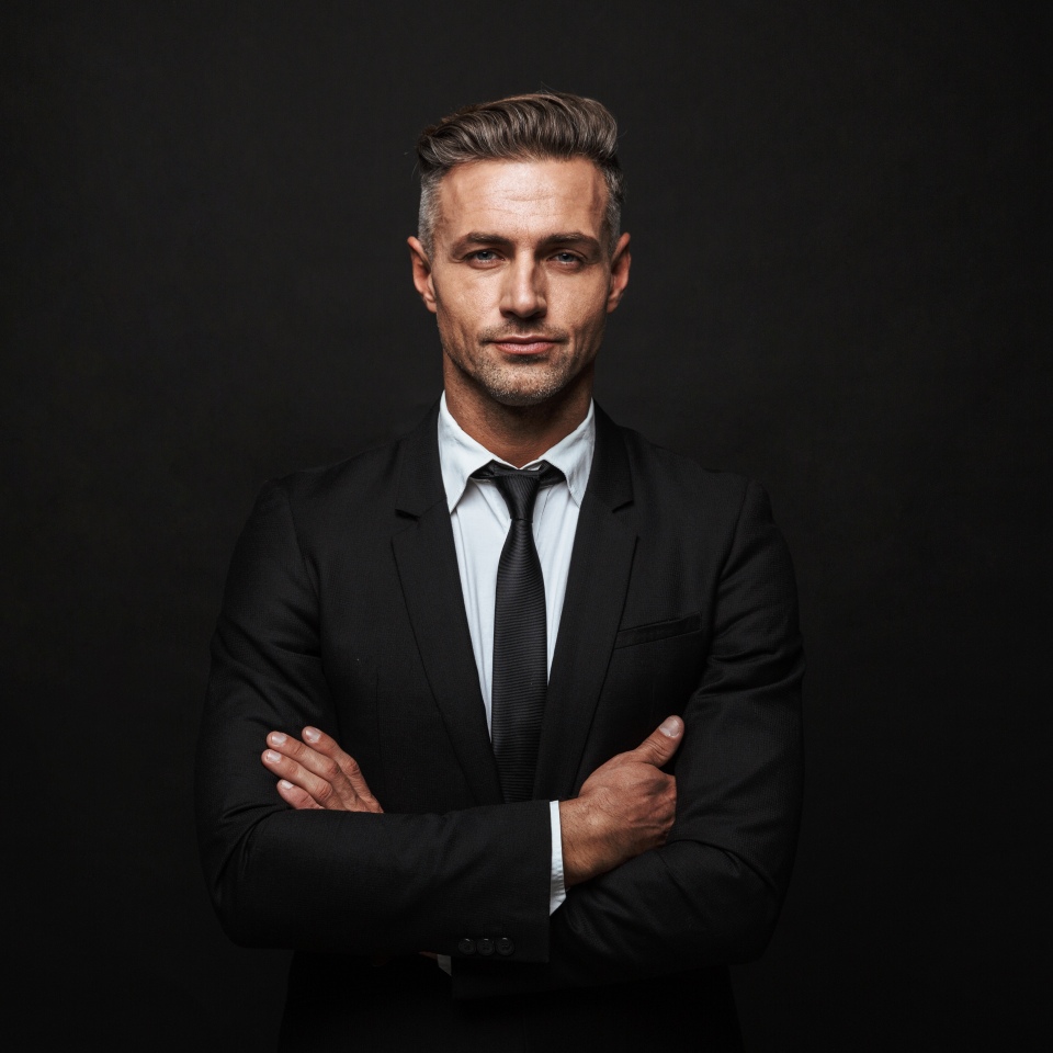 A person in a black suit and tie stands with arms crossed against a dark background.