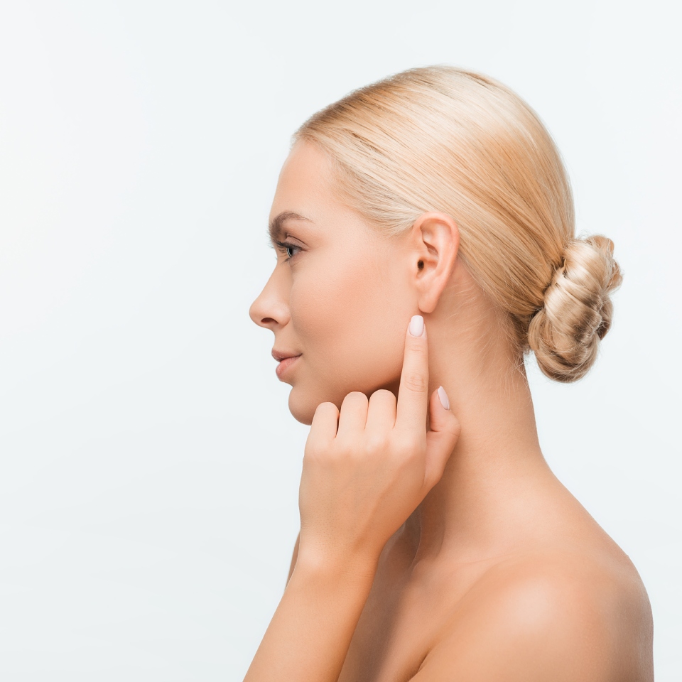 Woman with blonde hair in a bun pointing to her ear, side profile on a light background.