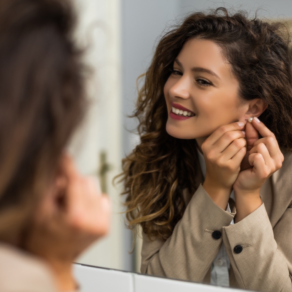 A woman with curly hair smiles while putting on an earring in front of a mirror.