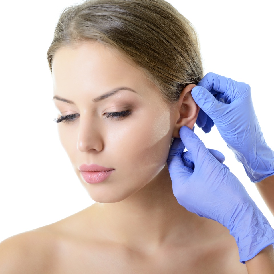 A person wearing blue gloves adjusts the earlobe of a woman with light skin and brown hair, against a white background.