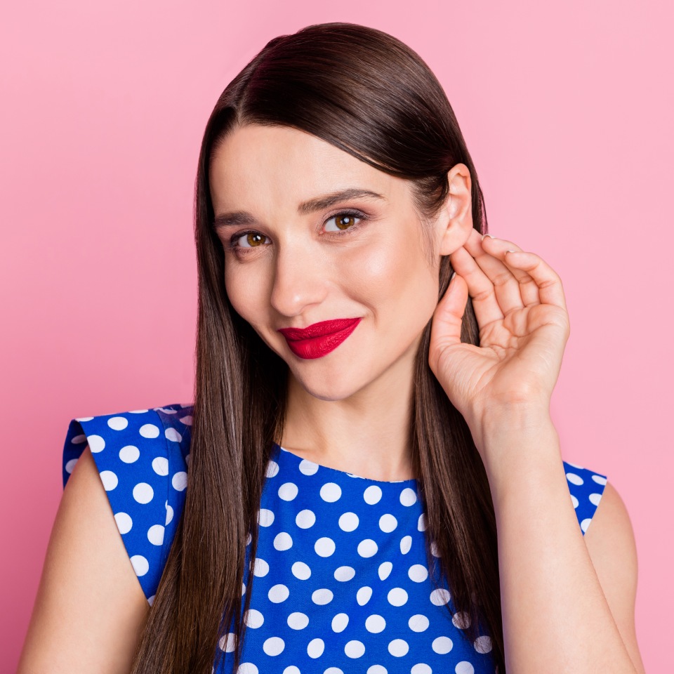 A woman with long brown hair in a blue polka dot dress holds her hand to her ear, smiling, against a pink background.