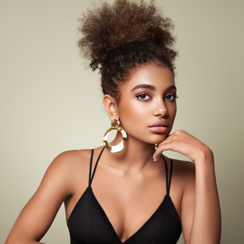 A woman with curly hair wears large gold earrings and a black sleeveless top, posing with her hand under her chin against a light green background.