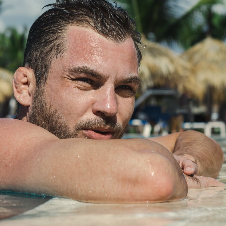 Man leaning on the edge of a swimming pool, with palm trees and thatched umbrellas in the background.