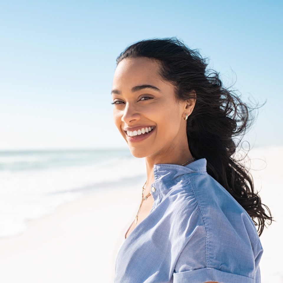 A person with dark hair smiles at the camera while standing on a sunny beach. They wear a light blue shirt and the ocean is visible in the background.