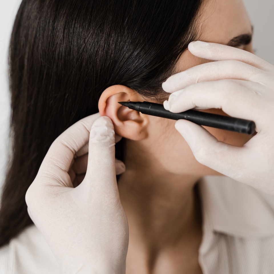 A person with dark hair receives a small black pen marking on their ear by gloved hands.