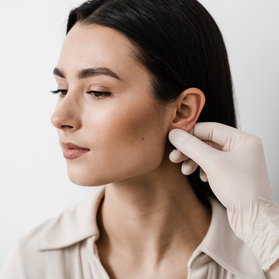 A person with long dark hair looks to the side while a gloved hand examines their ear.