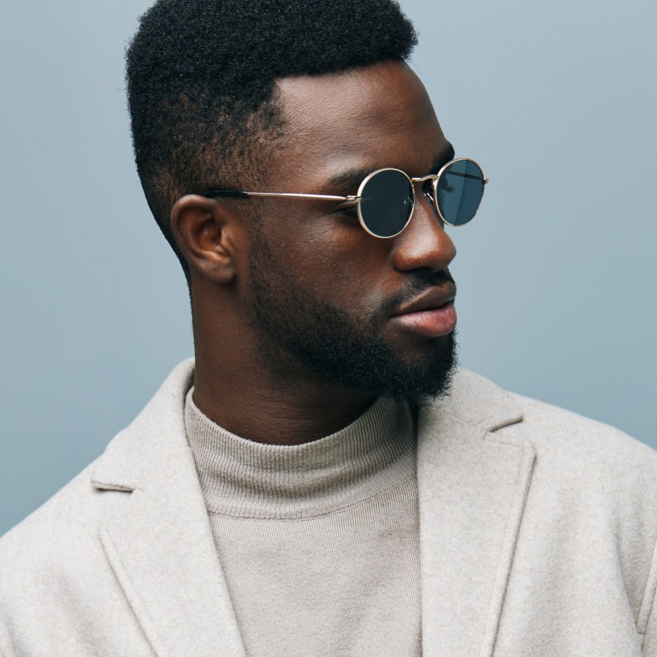 Man wearing round sunglasses and a light gray coat against a plain background, looking to the side.