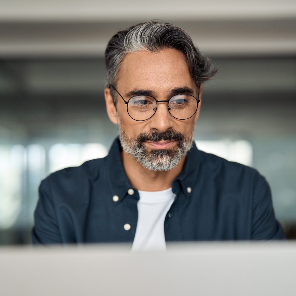 Man with glasses and gray beard looking at a computer screen, wearing a dark shirt over a white t-shirt.