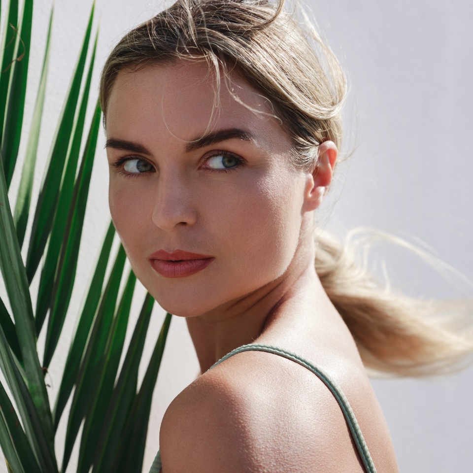 Woman with blonde hair and a green top stands in front of a white wall, holding a palm leaf.