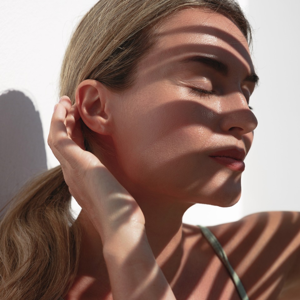 A person with eyes closed and hand near their face stands in dappled sunlight casting leaf shadows on their skin.