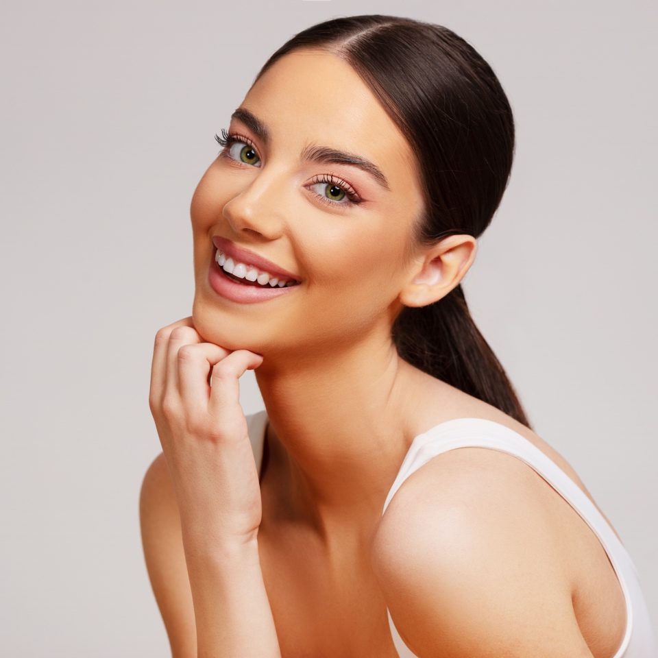 A woman with long dark hair in a ponytail smiles at the camera, resting her chin on her hand. She is wearing a white top against a plain background.