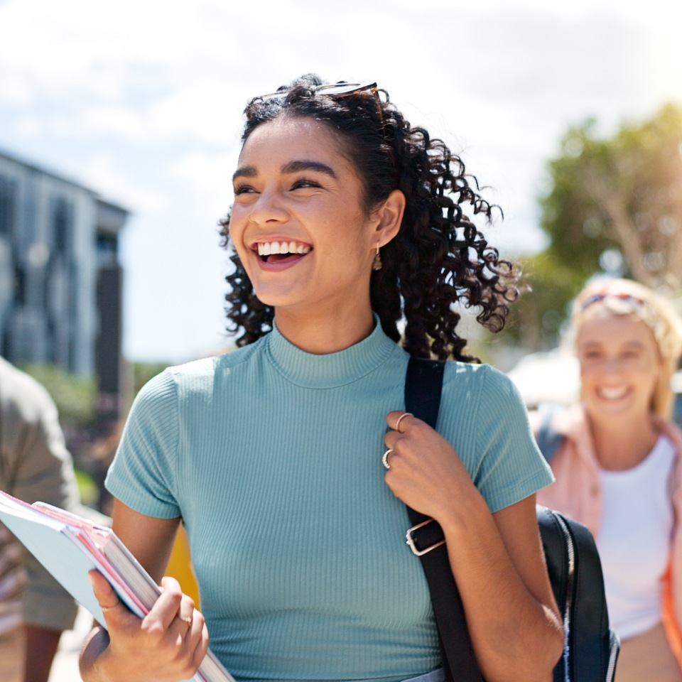 A person wearing a blue shirt smiles while holding notebooks. They are walking outside along with other people in the background.