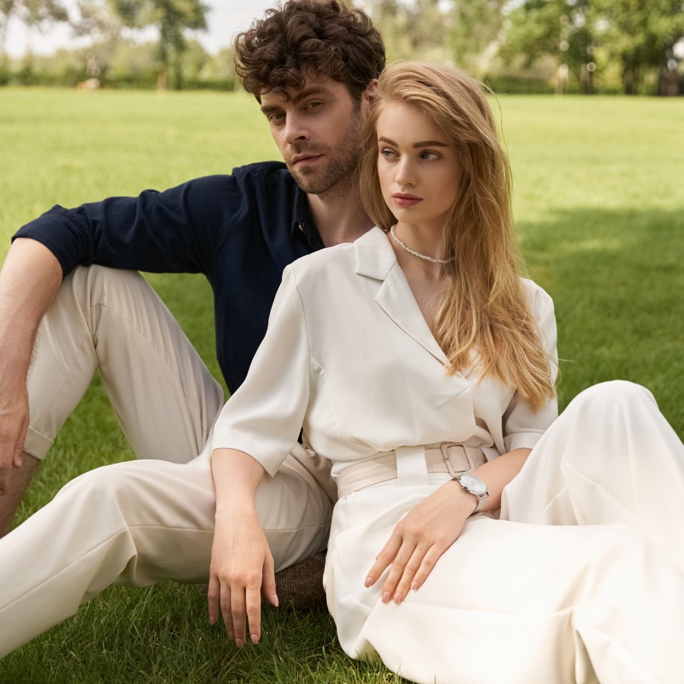 A man and woman sit close together on the grass in a park. The woman wears a white outfit and a watch, while the man wears a dark shirt and light pants. Trees are visible in the background.