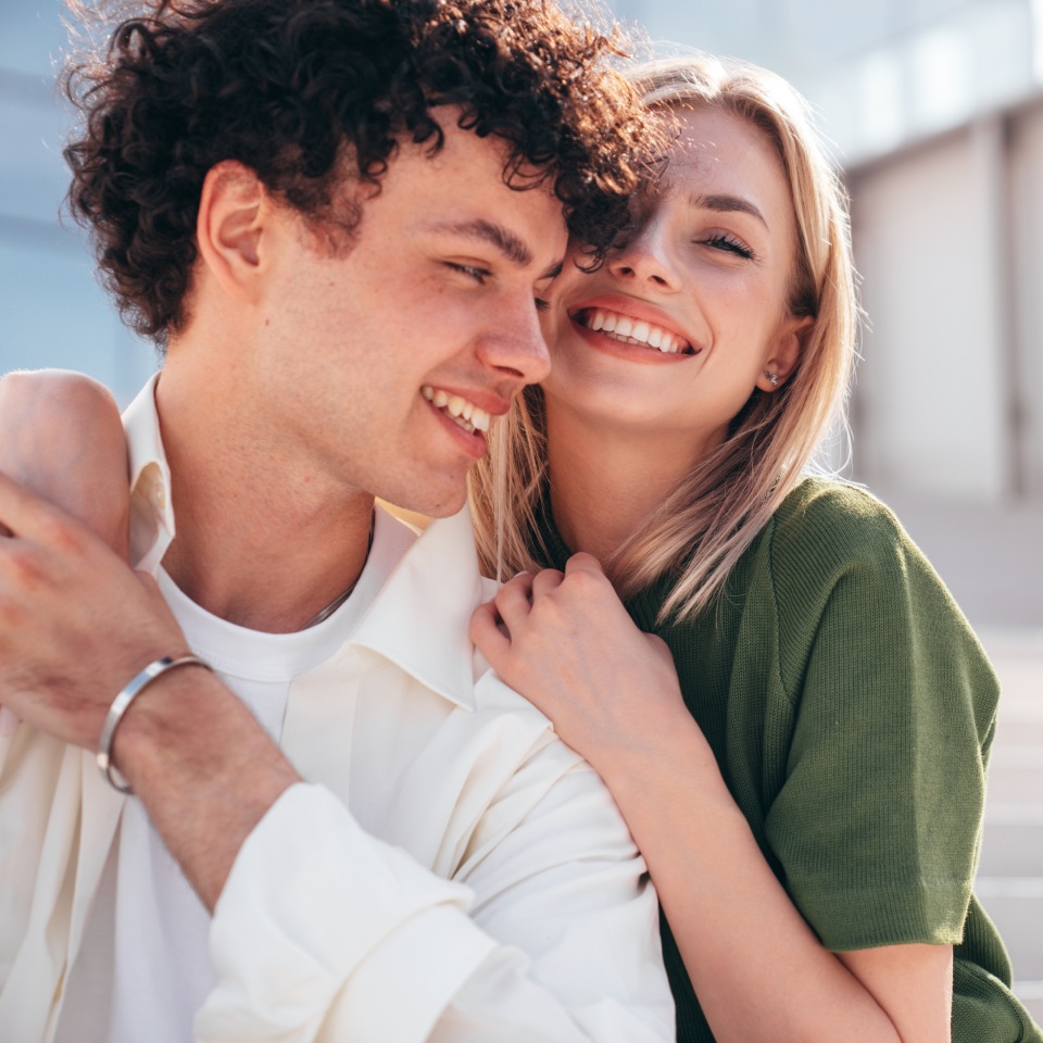 A man and woman smiling while sitting closely together outdoors. The woman leans on the man's shoulder, and both look cheerful.